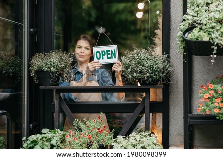 Small business owner smiling while turning sign for reopening of place after quarantine due to covid-19. Young lady in apron with nameplate on glass door of studio, flower shop, restaurant or eco cafe