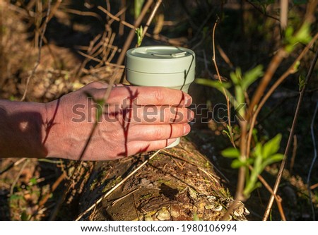 Reusable silicon coffee cup in forest taken by male hand among green plants, trees. Eco lifestyle. Beautiful day light and shadows.