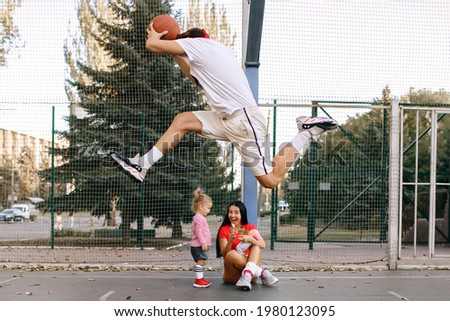 An athletic family spends the evening on the basketball court playing sports. Little girl and her mom are having fun while dad is jumping with a basket ball. Young parents dressed in sportswear