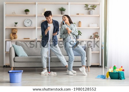 Funny asian loving couple singing songs while cleaning apartment, using broom and mop as microphones, cheerful beautiful young man and woman imitating rock stars while house-keeping, copy space Royalty-Free Stock Photo #1980128552