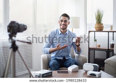 Arab male blogger promoting new model of wireless earphones, filming video review of modern gadget at home. Famous influencer making content for his tech channel, recommending innovative product Royalty-Free Stock Photo #1980128633