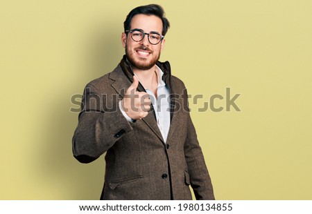 Young hispanic man wearing business jacket and glasses doing happy thumbs up gesture with hand. approving expression looking at the camera showing success. 
