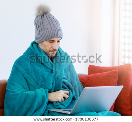 Caucasian unwell sick unhappy male employee wears knitted hat cover by warm blanket take day off from fever sit on leather couch working from home with laptop computer in freezing cold weather winter. Royalty-Free Stock Photo #1980138377