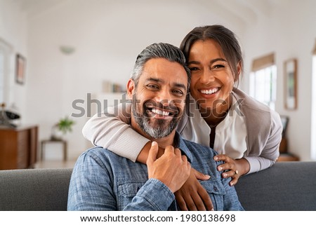 Portrait of multiethnic couple embracing and looking at camera sitting on sofa. Smiling african american woman hugging mid adult man sitting on couch from behind at home. Happy mixed race couple laugh Royalty-Free Stock Photo #1980138698