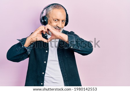 Handsome senior man with beard listening to music using headphones smiling in love doing heart symbol shape with hands. romantic concept. 