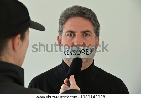 Censored eyewitness attractive adult person man, (male age 40-50) with mouth shut with tape, being interviewed by fake news reporter.Freedom of the press and media concept. Real people. Copy space Royalty-Free Stock Photo #1980145058
