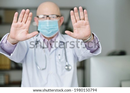 Portrait of bald mature doctor in lab coat standing in office and making stop sign