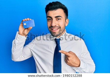 Young hispanic man wearing business clothes holding credit card smiling happy pointing with hand and finger 