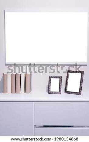 A white chest of drawers on which there are two frames for photographs and several books in beige and white colors. There is a large mirror on the wall.
