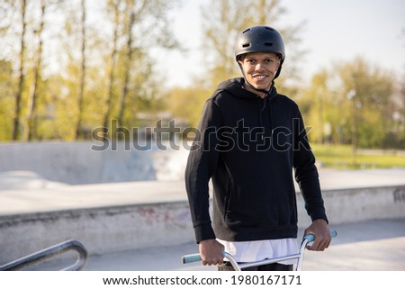 A teenager in a black sweatshirt and helmet stands on a ramp at a skatepark and smiles for the camera, his face illuminated by the setting sun