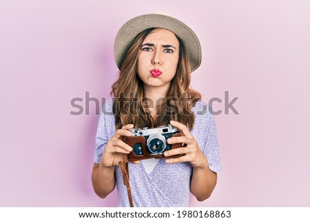 Young hispanic girl wearing summer hat holding vintage camera puffing cheeks with funny face. mouth inflated with air, catching air. 