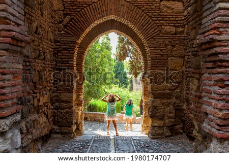 Gateway of the Alcazaba, Moorish fort in the old center of Malaga, Spain. Two girls discover the historic monument with old walls and green trees.  Royalty-Free Stock Photo #1980174707
