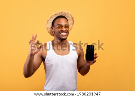 Awesome mobile application. Carefree African American guy in summer wear holding smartphone with mockup for website or app on screen, showing okay gesture over yellow studio background