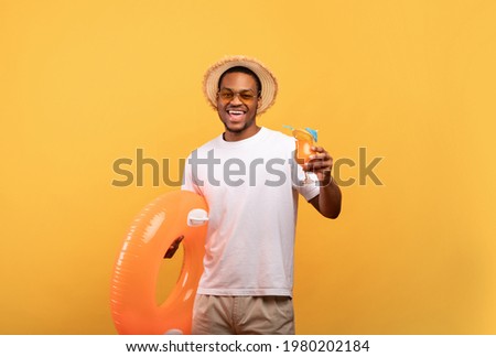 Joyful black man with tasty summer cocktail holding inflatable ring, going to beach party on yellow studio background. Attractive African American guy with fresh summertime drink ready to chill