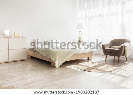 Simple design, ad, offer and modern home style. Double bed with pillows, soft blanket, carpet, lamp, armchair and furniture, on wooden floor. White wall, big window with curtains in bedroom interior Royalty-Free Stock Photo #1980202187
