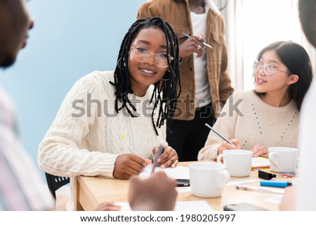 Woman in glasses with dark complexion and pigtails sits in office at work in conference meeting with colleagues, discussing business project, filling out documents, drinking coffee, corporate work