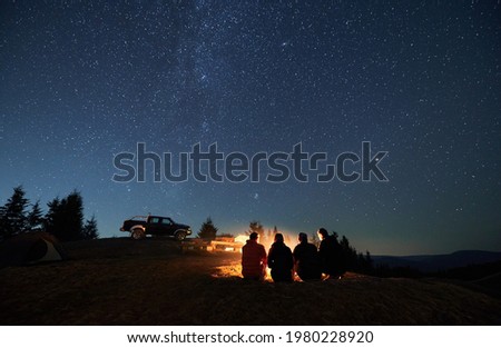 Evening starry sky over mountain valley with car and hikers near campfire. Group of travelers sitting near bonfire under majestic blue sky with stars. Concept of night camping, hiking and travelling. Royalty-Free Stock Photo #1980228920