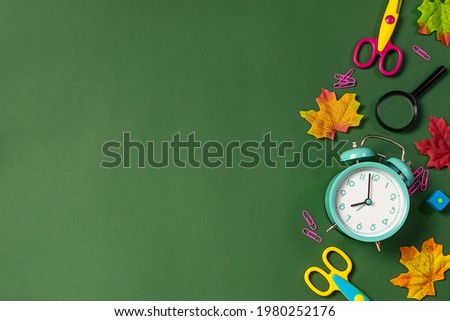 Back to school background concept with bright stationery, alarm clock and autumn leaves on deep green background with copy space for your design.