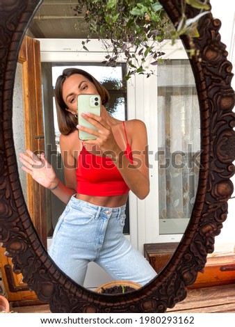 Pretty woman at home take photo selfie in mirror on mobile phone for stories and posts in social media, vertical frame, wearing jeans and red top, fit slender, good looking