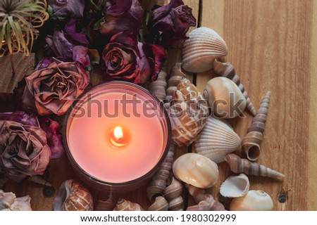 Close-up image of a burning pink candle surrounded by seashells, dried pink roses on a wooden surface
