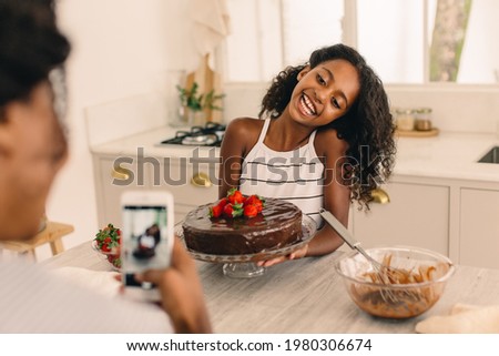 Girl with freshly baked cake being photographed by mother in kitchen. Mother taking picture woman daughter making cake.