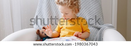 Cute baby boy toddler sitting at kids room and reading a book. Early age kid child development literacy alphabet education. Candid home authentic childhood lifestyle. Web banner header.