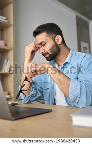 Young indian exhausted business man rubbing nose bridge in modern home office with laptop on desk. Overworked burnout academic Hispanic student with glasses in hand feeling eyestrain. Vertical shot. Royalty-Free Stock Photo #1980428450