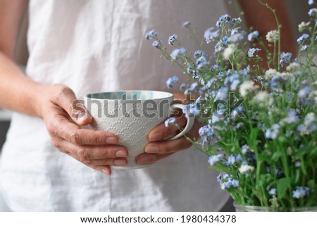 Woman's hands holding a white cup of tea or coffee beside blue flowers Royalty-Free Stock Photo #1980434378