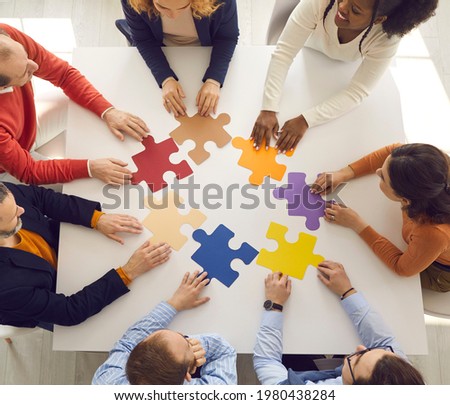 Team of young and senior employees sitting in circle around office table putting jigsaw puzzle together as metaphor for teamwork and looking for working business solutions. Square format, high angle Royalty-Free Stock Photo #1980438284