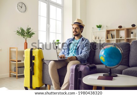 Young man planning vacation trip and using computer to book hotel room online. Happy male tourist wearing fedora hat sitting on sofa at home and browsing rental holiday accommodation website on laptop Royalty-Free Stock Photo #1980438320