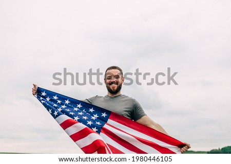 Happy smiling man in military color t-shirt holding in hands big American flag sincerely rejoicing. Green field on the background. Patriotism