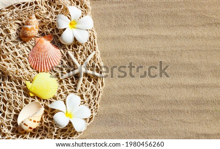 White plumeria flowers with starfish and seashells on mesh beach bag on sand. Top view