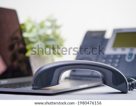 In the photo, the office phone receiver is on a laptop. In the background is an indoor flower in a white pot. Gray and white tones. Close-up. There are no people in the photo.