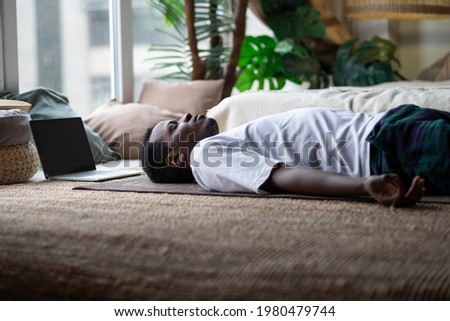 Yoga. African young man meditating on a floor and lying in Shavasana pose. Royalty-Free Stock Photo #1980479744