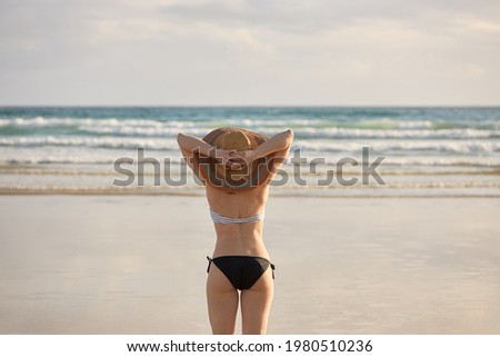 Girl in bikini with straw hat on the shore of a beach holding her straw hat.