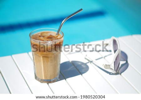 Ice coffee Cyprus Frappe Fredo against blue clear water of the swimming pool, on white table, with sunglasses. Summer minimalistic background, holiday or vacation concept. Sun and shadows.Copy space Royalty-Free Stock Photo #1980520304