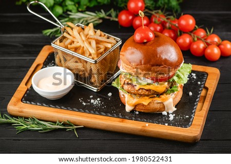 Side view of Cheeseburger with tomatoes, lettuce, pickled cucumber and sauce. Served with French fries and sauce on wooden board with ingredients on background.