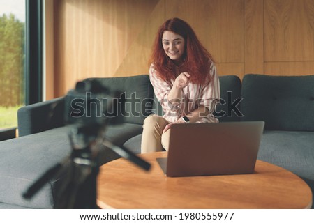 Young redhead freelancer woman recording video with camera, tripod, laptop. Smiling woman sitting at modern house, looking at laptop screen, finger points at laptop while recording online webinar