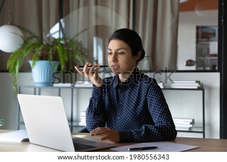 Serious young Indian businesswoman work online on computer in office record audio voice message on smartphone. Focused ethnic female employee buys with laptop talk on loudspeaker on cellphone.