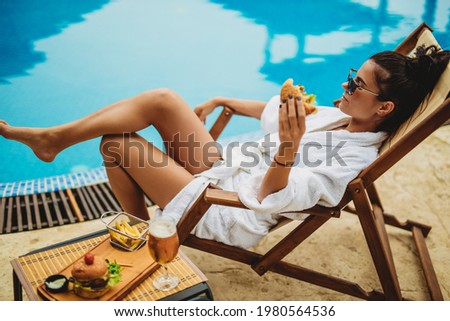 Young attractive woman holding a burger in her hand and sitting by the pool and chilling stock photo. Close-up shot.