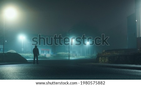 A mysterious hooded man standing in a street on an industrial estate looking at buildings on a misty winters night.         Royalty-Free Stock Photo #1980575882