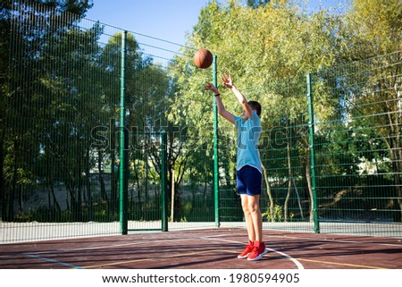 Cute boy in green t shirt plays basketball on a city playground. Active teen enjoying outdoor game with orange ball. Hobby, active lifestyle, sport for kids.	
