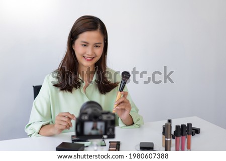 A beauty influencer applying powder blush on her cheek using makeup brush in front of a camera.