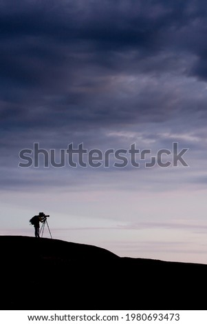 Side view of a young man taking pictures with camera and telephoto zoom lens on tripod on the mountain peak at sunset. Dramatic dark rain clouds in the backgrounds. Travel exploration concepts.