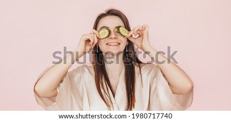 Girl make homemade face beauty masks. Cucumbers for the freshness of the skin around the eyes. Woman take care of youthful skin. Model laughing and having fun in spa on pink background