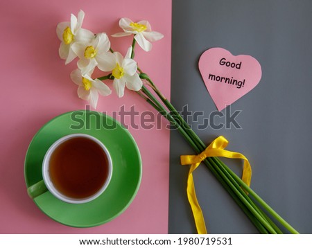 Good morning wishes note in shape heart, tea cup and white yellow daffodils bouquet on bicolor pink and grey paper trend minimalism background top view flat lay horizontal