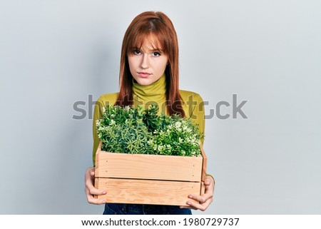 Redhead young woman holding wooden plant pot relaxed with serious expression on face. simple and natural looking at the camera. 