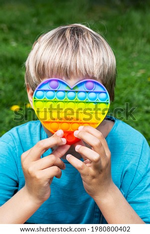 Child plays on the grass with a simple dimple relaxing  toy. Little boy playing with pop it toy heart shaped, rainbow colored at summer day.