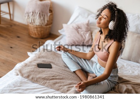 Smiling mid aged african woman listening to music with headphones while relaxing on bed at home, meditating Royalty-Free Stock Photo #1980861341