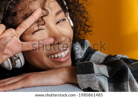 Young african woman in headphones winking and showing peace sign isolated over brown wall